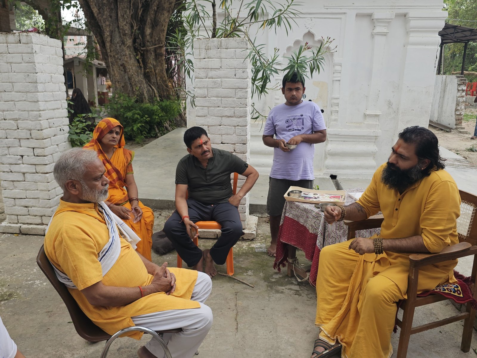 Kabeer Ji Maharaj मंदिर परिसर में भक्तों के बीच discourse — एक महिला, पुरुष और युवा के साथ। मंदिर की दीवारें, हरा वातावरण।