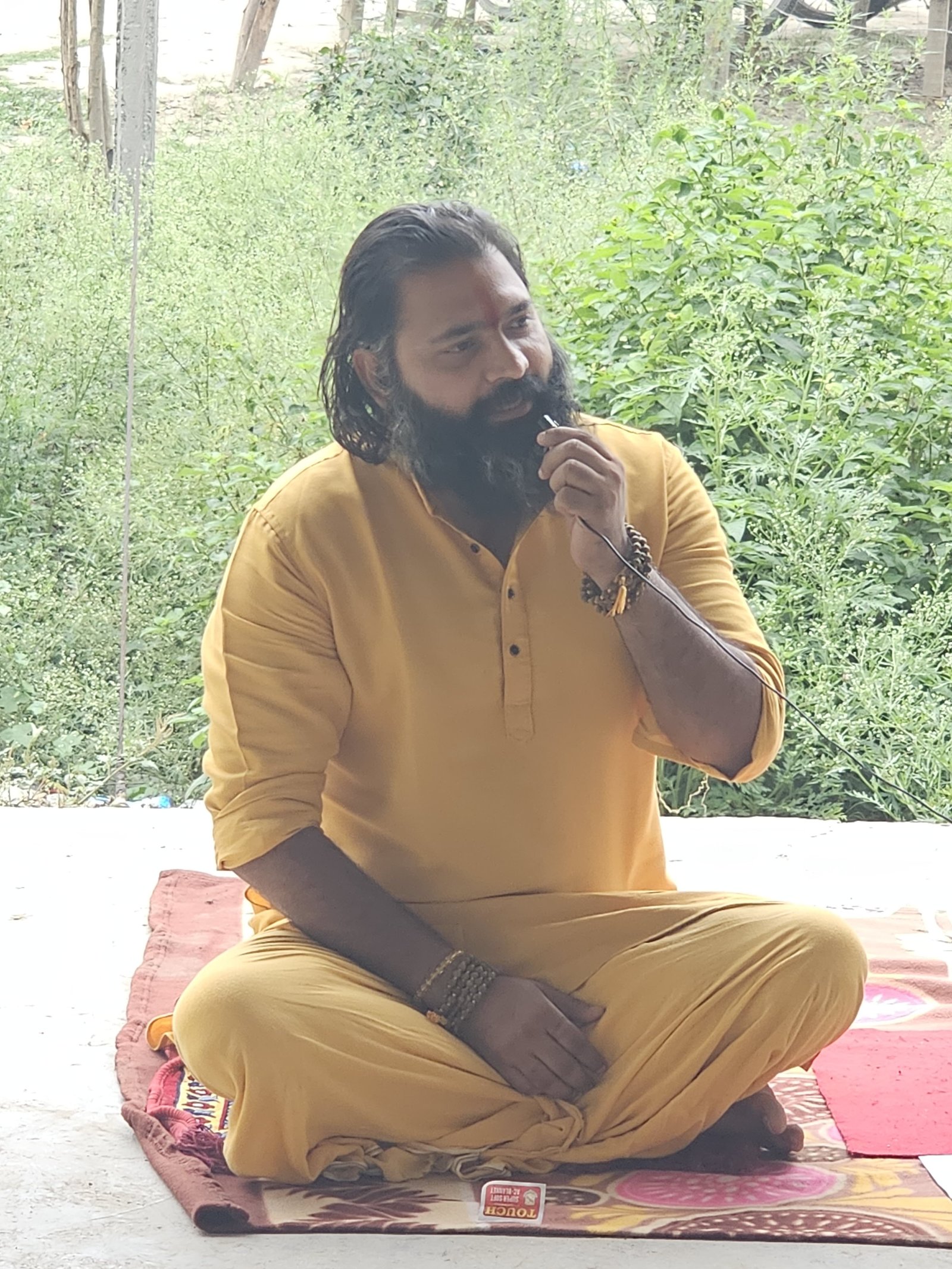 Kabeer Ji Maharaj प्रकृति के बीच ध्यानमग्न मुद्रा में बैठे हैं — भगवा वस्त्र, रुद्राक्ष, हरियाली। Daily Discourse session।
