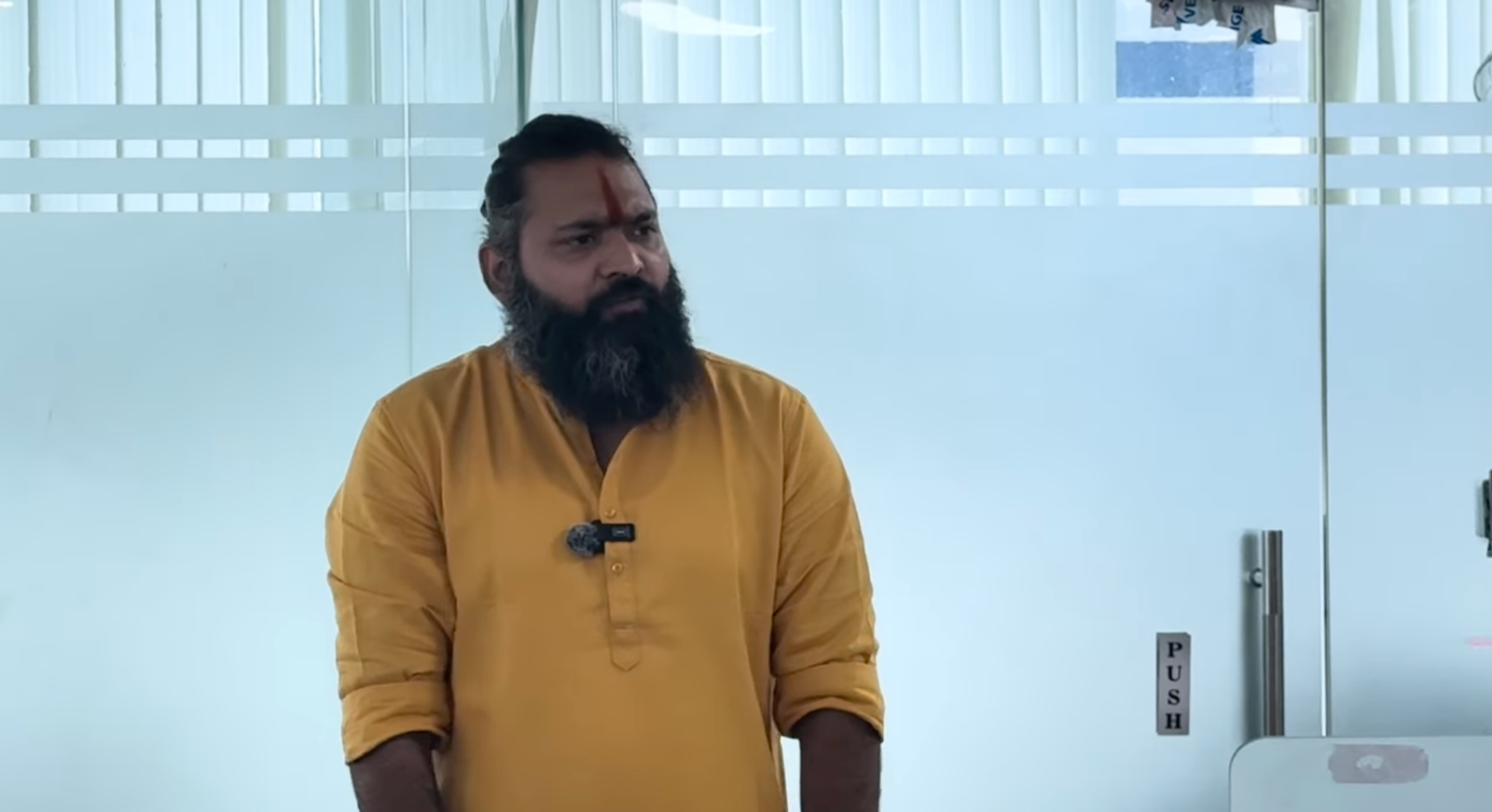 Kabeer Goswami corporate office में close-up — भगवा कुर्ता, तिलक, दाढ़ी — thoughtful expression। Glass partition पृष्ठभूमि।