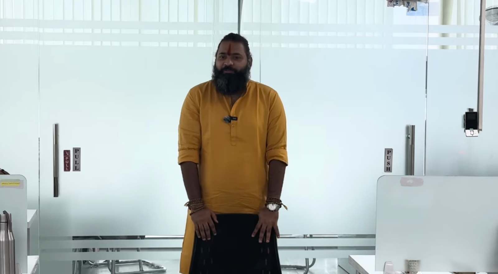 Kabeer Goswami office glass doors के बीच खड़े — पूरा frame। भगवा कुर्ता, तिलक, PUSH door handle। Corporate और spiritual का अनोखा संगम।