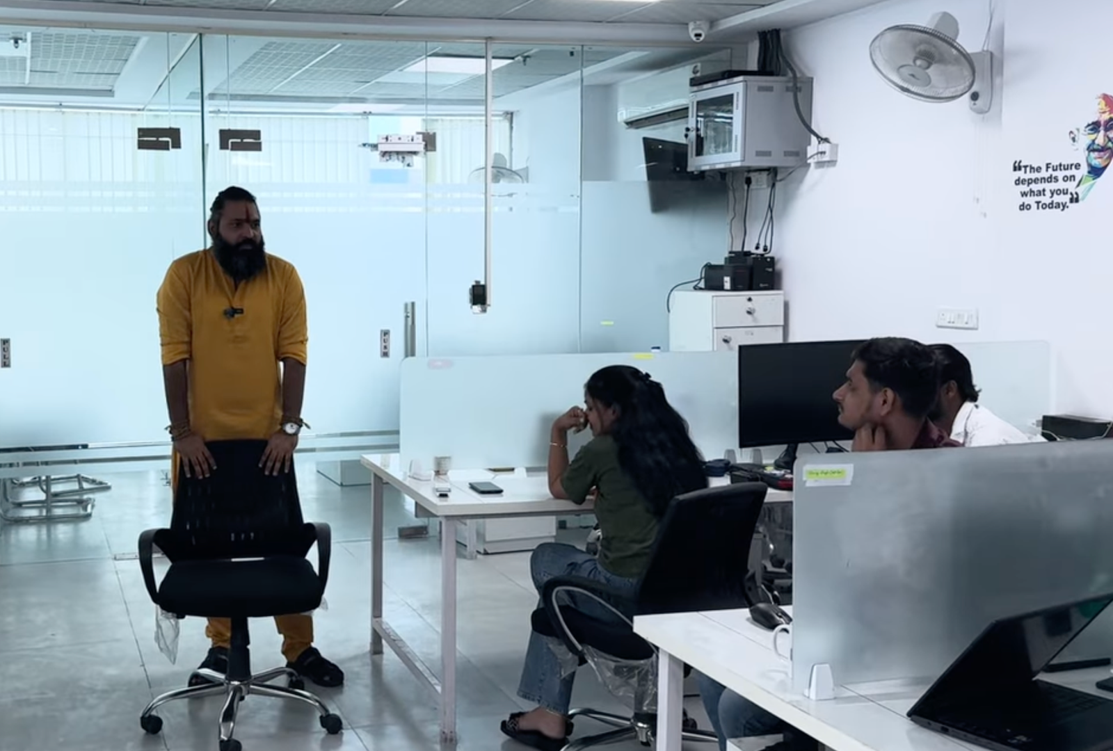 Kabeer Goswami (Kabeer Ji Maharaj) सफेद modern corporate office में खड़े होकर team को address कर रहे हैं। भगवा कुर्ता, पृष्ठभूमि में Gandhi quote — The Future depends on what you do Today.